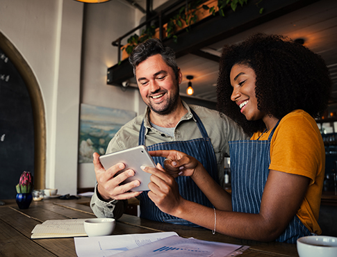 man and woman that work in a coffee shop wearing aprons looking at ipad together